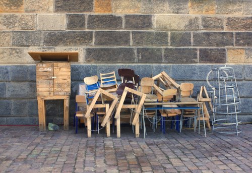 A collection of assorted wooden chairs and small tables, some stacked and leaning against each other, is positioned on a cobbled pavement in front of a weathered brick wall. The chairs and tables vary in design, with some featuring natural wood finishes, while others are painted in shades of beige and dark brown. To the left, there is a small wooden cabinet or storage unit with a slatted door, also made of natural wood, standing against the brick wall. On the right side, a metal wheeled cart or trolley with a wire frame is visible, leaning slightly against the wall. The scene appears to be an outdoor area, possibly in a commercial or residential space, with the objects likely discarded or awaiting collection, which aligns with rubbish removal services in Thamesmead. The lighting suggests daylight, and the overall impression is of a cluttered outdoor space with a variety of wooden and metal objects, demonstrating typical rubbish items for removal service by Garden Clearance Thamesmead.