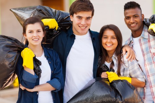 In the foreground, three young adults and one teenager are standing outdoors in a residential area, holding large black rubbish bags filled with waste, indicating participation in a rubbish removal activity. The individuals are dressed casually, with some wearing yellow cleaning gloves, and they are smiling at the camera, suggesting a positive community effort. The background shows a blurred urban setting with a building and pavement, typical of a Thamesmead neighborhood, subtly indicating a local context. The scene reflects a cleanup or garden clearance activity, and the group’s engagement aligns with services provided by Garden Clearance Thamesmead, specializing in rubbish removal and clearance tasks. Natural daylight illuminates the scene, highlighting the textures of the rubbish bags, the casual clothing's fabric, and the cheerful expressions of the participants, conveying a sense of community involvement and environmental care.