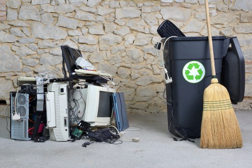A pile of discarded electronic waste, including computer monitors, keyboards, and tower cases, is stacked on a concrete floor next to a large black rubbish bin with a green recycling symbol in front of a textured stone wall. The electronic items appear to be secondhand or broken components, with some cables and small parts visible among them. To the right of the bin, there is a yellow straw broom leaning against it. The scene suggests an outdoor or garage area set in Thamesmead, where Garden Clearance Thamesmead offers rubbish removal services, highlighting proper disposal of electronic waste alongside general rubbish collection. The overall environment is clean, and the setup emphasizes responsible waste management practices related to household or garden clearance, suited for local areas like Thamesmead or surrounding regions in South East London.