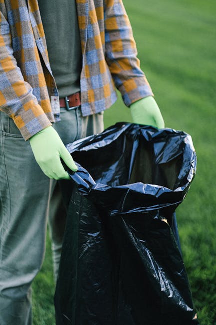 A person's arm extending from the left side of the image holding a green and yellow tied plastic rubbish bag filled with waste, with some uneven creases and transparency revealing its contents. The ba